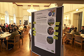 Poster zum Dialog Klimaanpassung im Vordergrund, dahinter ein Blick in den Saal mit den Teilnehmenden 